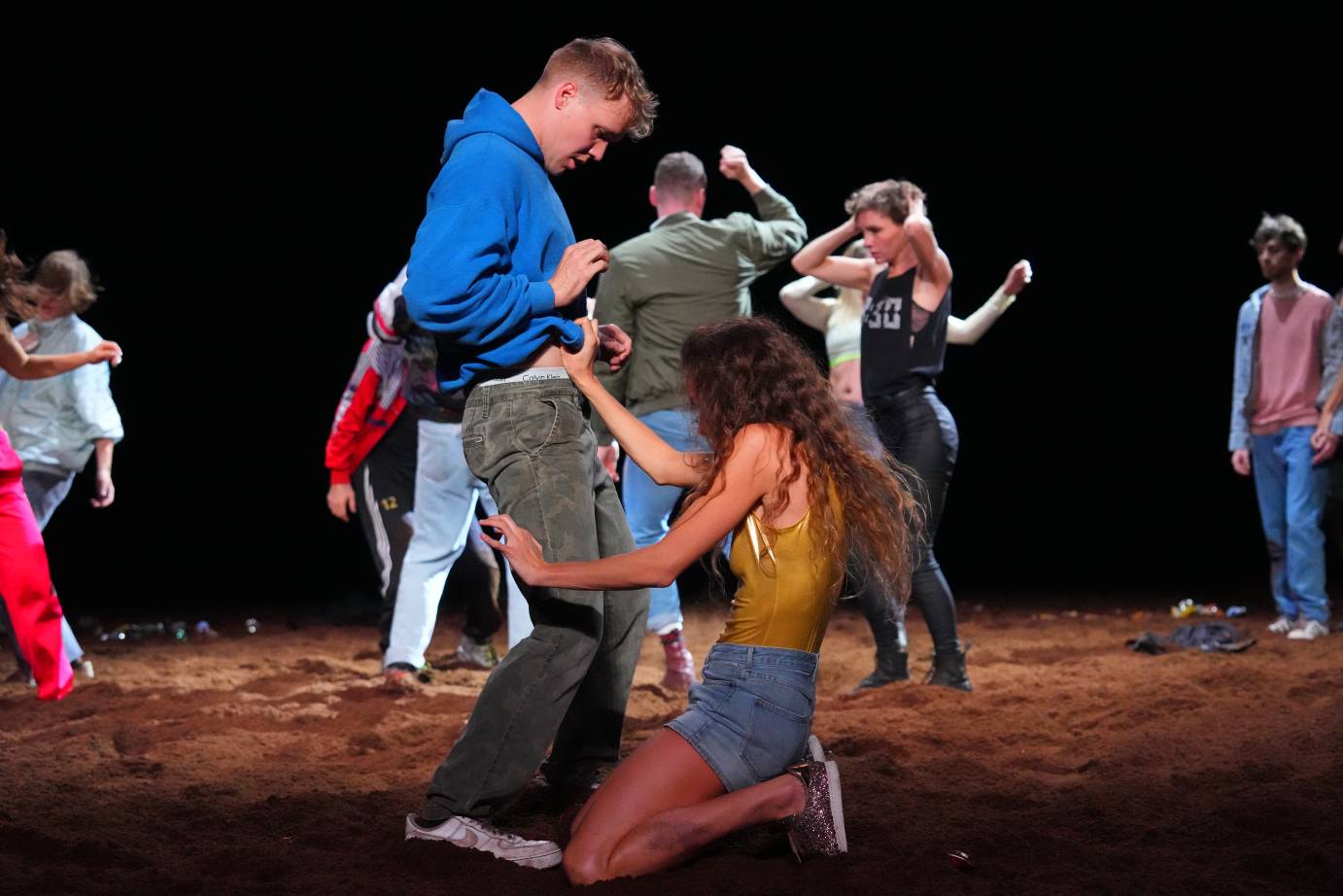 a woman in a golden leotard and jean hot pants kneels in the dirt pulling at the jeans of a man who stands above her looking wasted